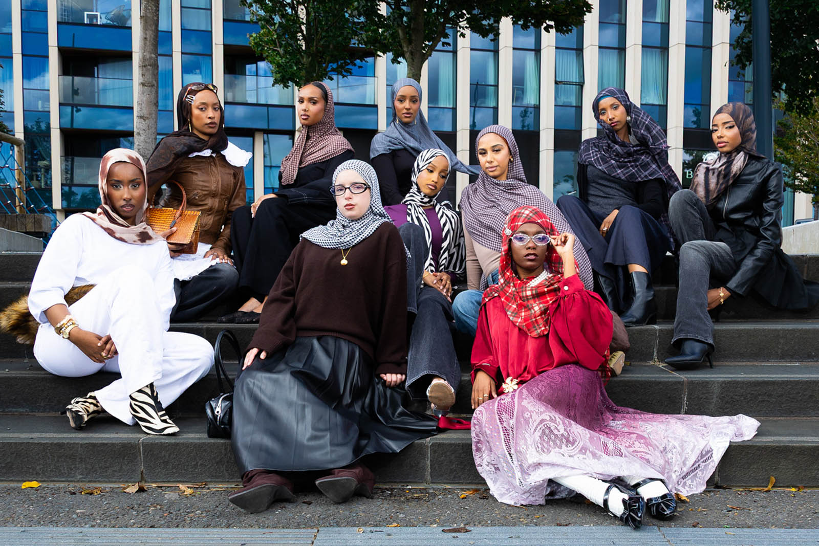 A photograph of a group of models, sitting on steps in what looks like a public square or plaza with an office block in the background, wearing hijabs by fashion brand Iftin