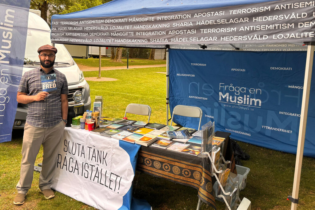 A photograph of Kashif Virk standing in front of the Fråga en Muslim (Ask A Muslim) stand at an event, wearing a flat cap and holding one of the organisation's leaflets