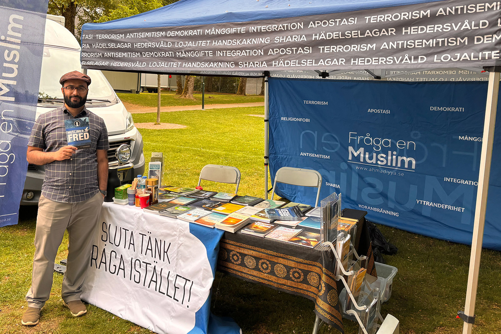 A photograph of Kashif Virk standing in front of the Fråga en Muslim (Ask A Muslim) stand at an event, wearing a flat cap and holding one of the organisation's leaflets