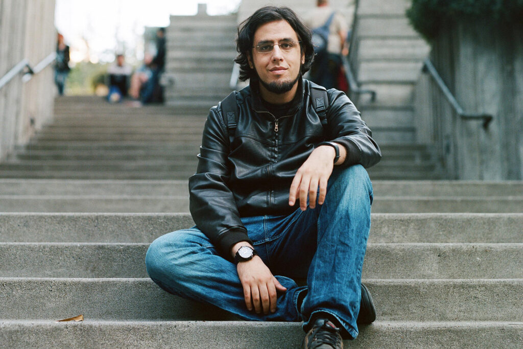 A portrait photograph of Rami Ismail, sitting on stone steps in jeans and a leather jacket, smiling at the camera
