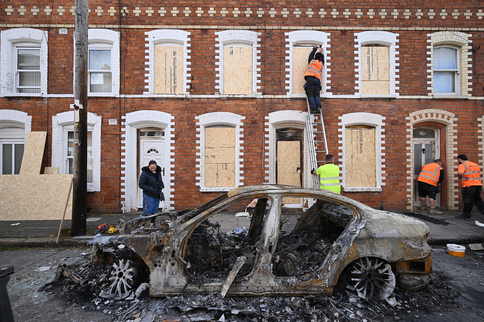 A terrace of houses with boarded up windows and a burnt out car in front of them. People in high-vis jackets examine the damage, with one up a ladder.