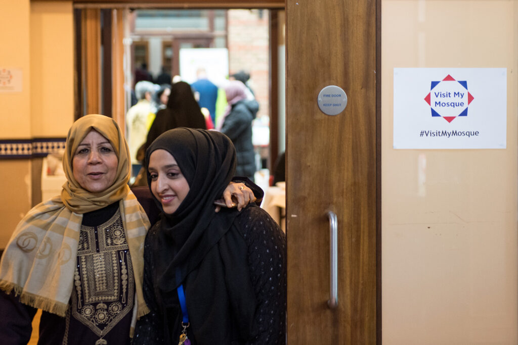 A photograph of two women wearing headscarves, one with her arm round the other, standing in a doorway during Visit My Mosque weekend at Al Manaar masjid, west London.