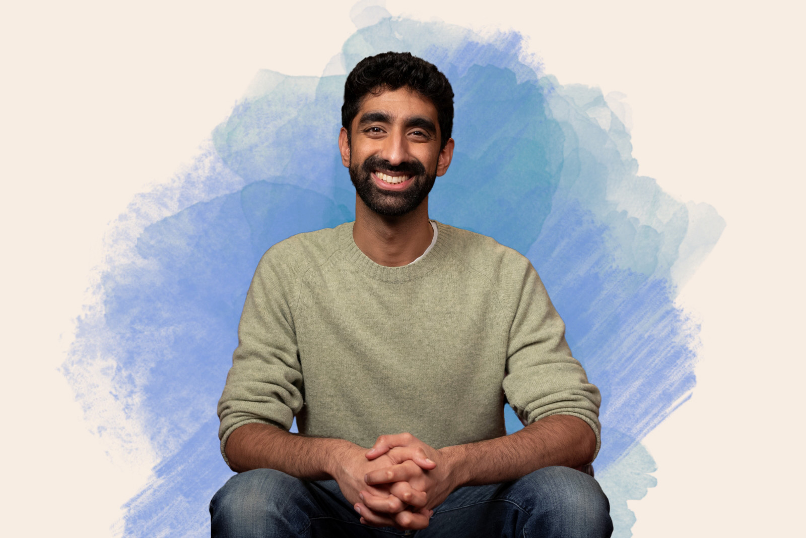 A portrait photograph of Kas Ahmed, TV doctor and Traitors faithful, seated wearing jeans and a jumper, smiling and with his hands clasped in front of him
