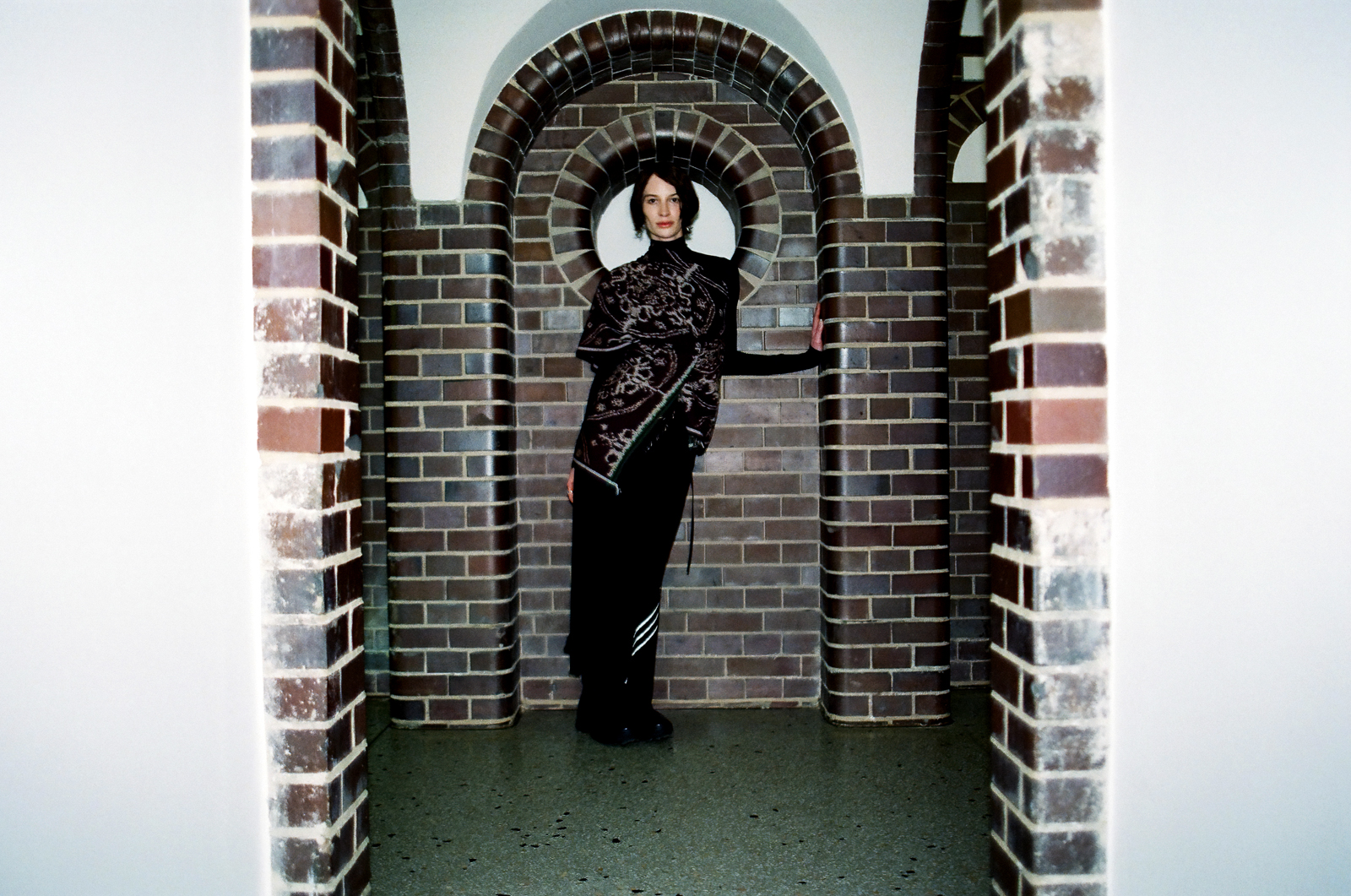 A photograph of Alif Hilal standing and leaning against a polished-brick wall in a corridor
