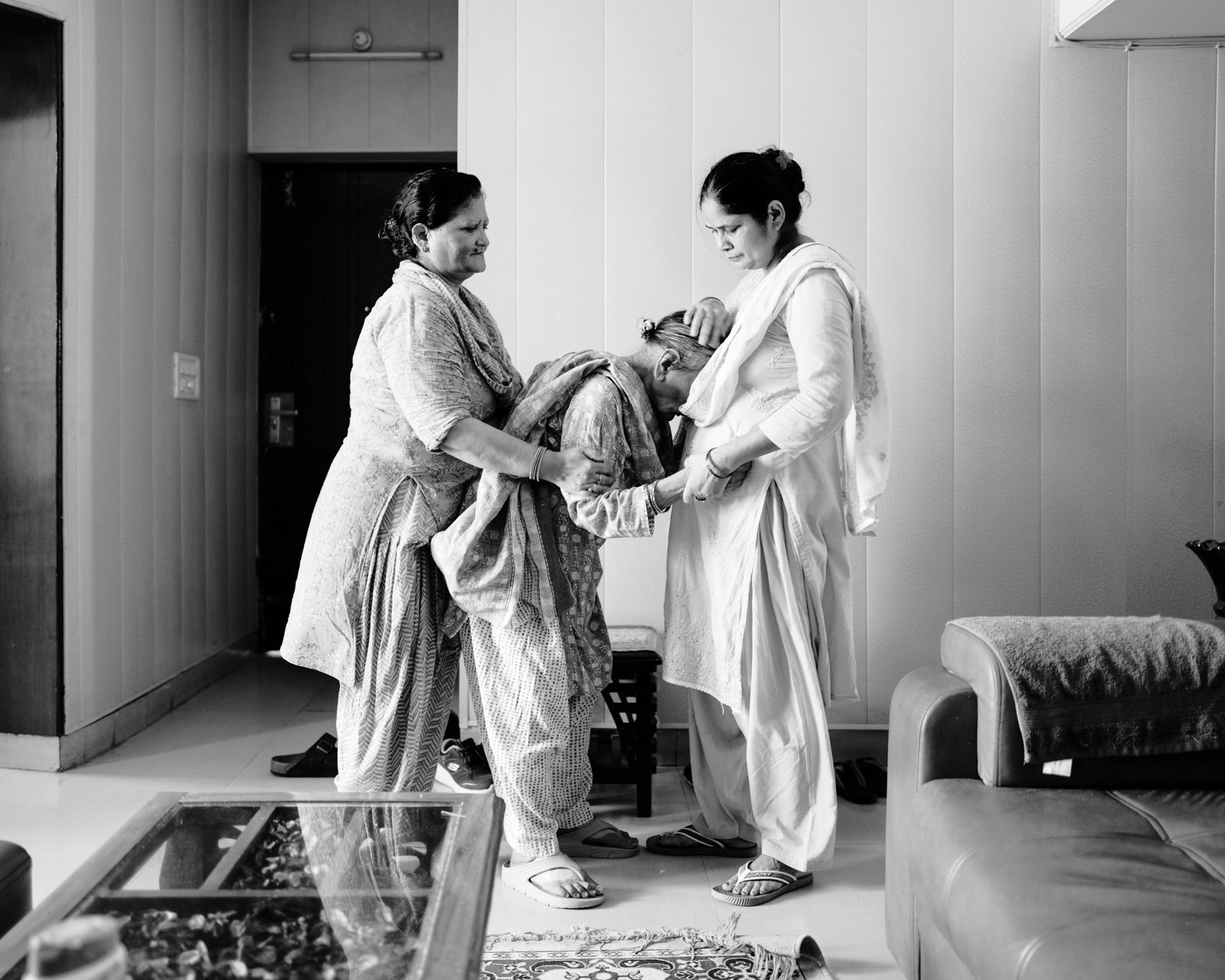 A black and white photograph from Aman Alam's series Ozymandias, documenting his grandmother's life with Alzheimer's, titled Naseem’s Morning Walk. In the image, Gudia and Laxmi, Naseem's two nurses, help her with her morning walk inside the house in Agra. She now needs help with the most basic of motor functions