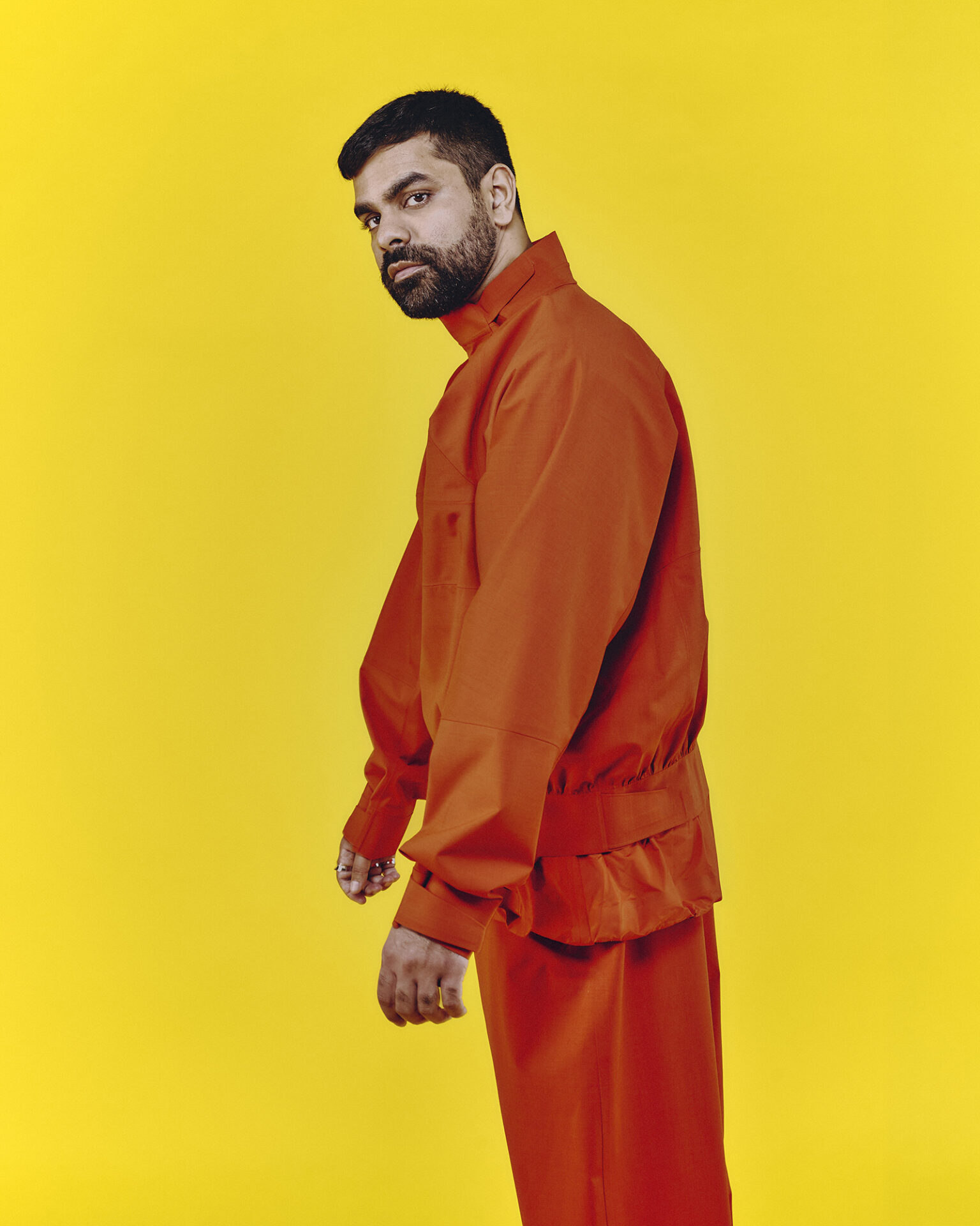 A portrait photograph of DJ and producer Ahadadream, sideways on to the camera wearing an orange outfit against a yellow background