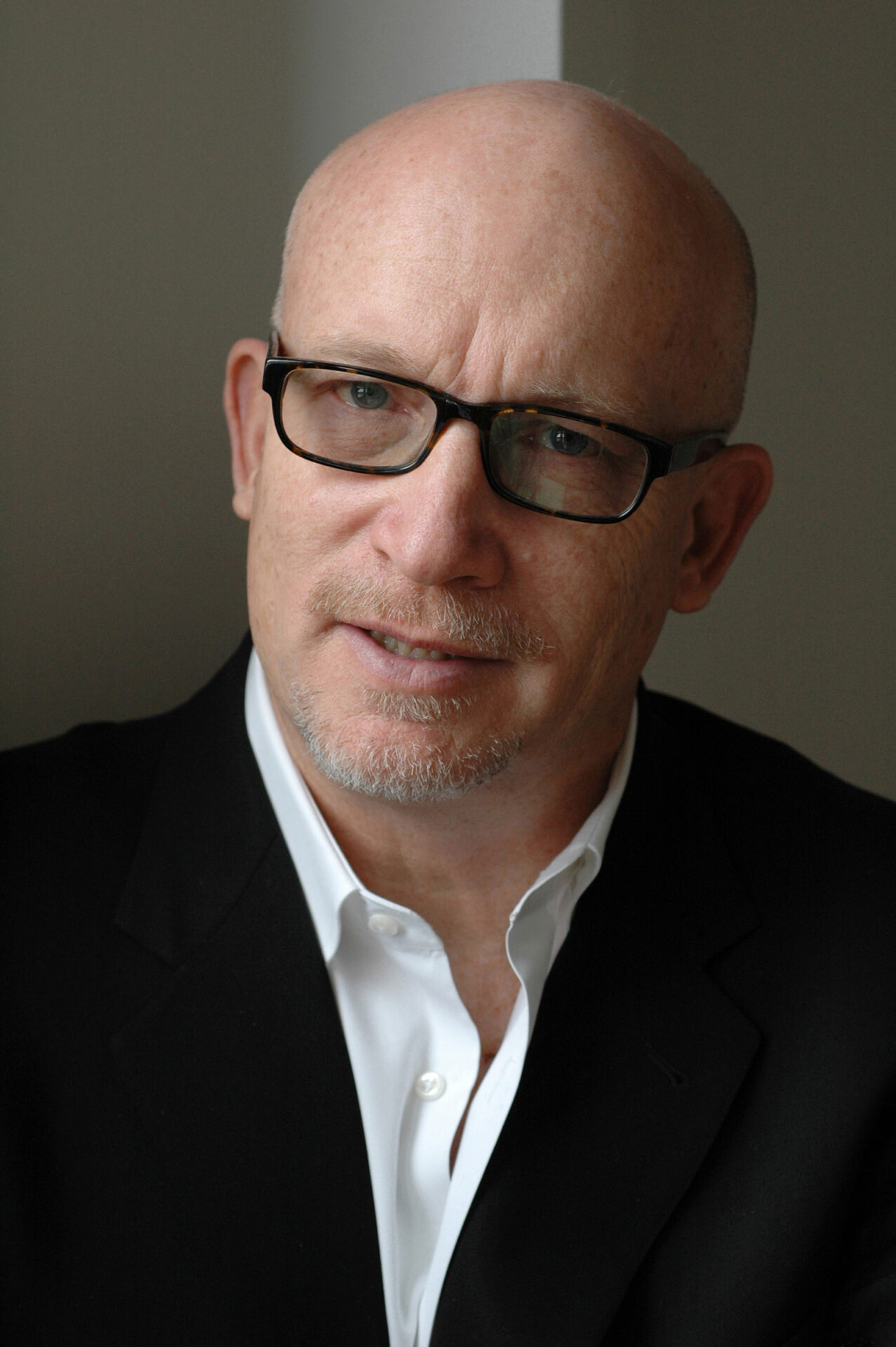 A head and shoulders portrait photograph of documentary-maker Alex Gibney, wearing glasses, an open-necked white collar shirt and black jacket