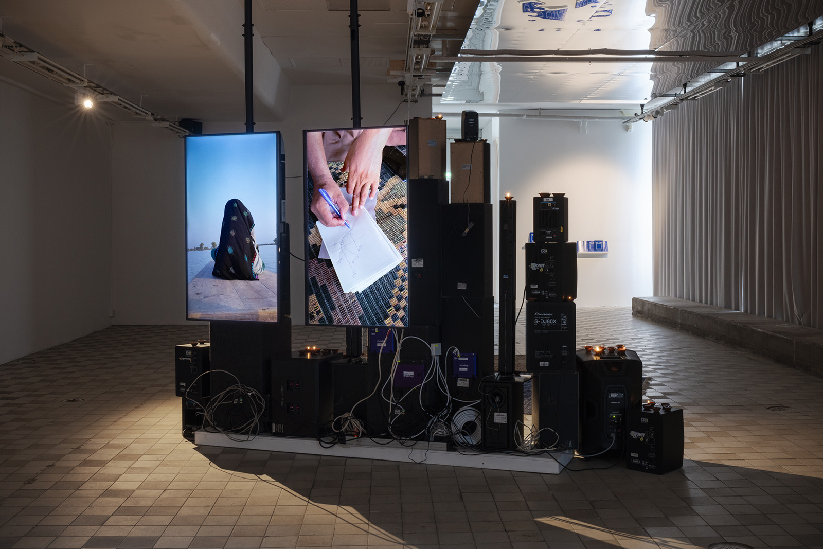 A photograph of Zahra Malkani's Sada Sada installation, part of Sacred Spaces at Stockholm's Konsthall C, featuring two large screens in the centre of a gallery surrounded by audiovisual equipment