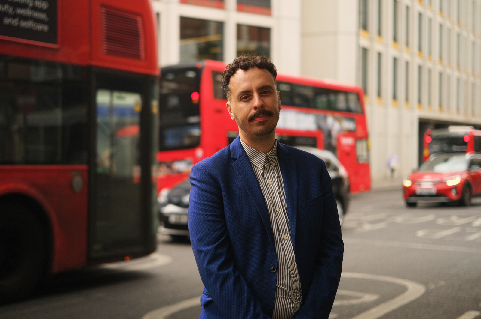 A photograph of Green Party London Assembly member Benali Hamdache, standing on the pavement with cars and red buses passing in the background