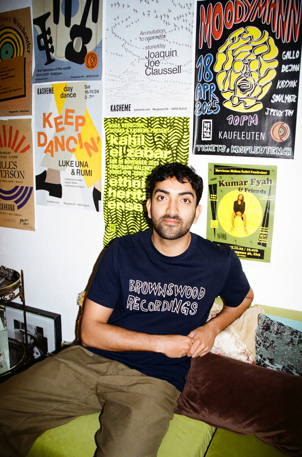 A photograph of Rohan Rakhit, the DJ, broadcaster and From The Margins co-curator, sitting in front of a wall with various posters for live music events