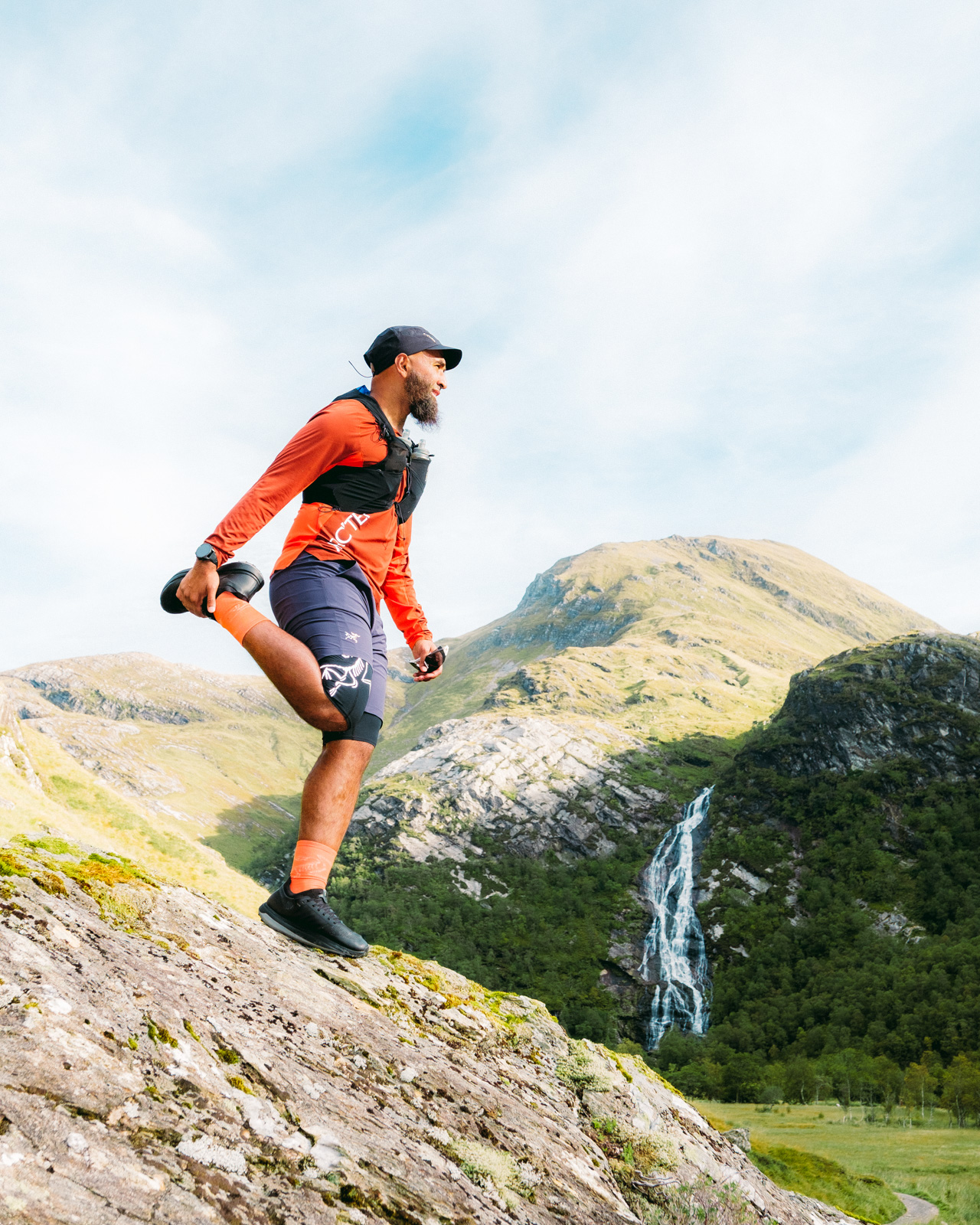 A photograph of a Muslim hiker, standing on a rock performing a leg stretch exercise, with Ben Nevis in the background