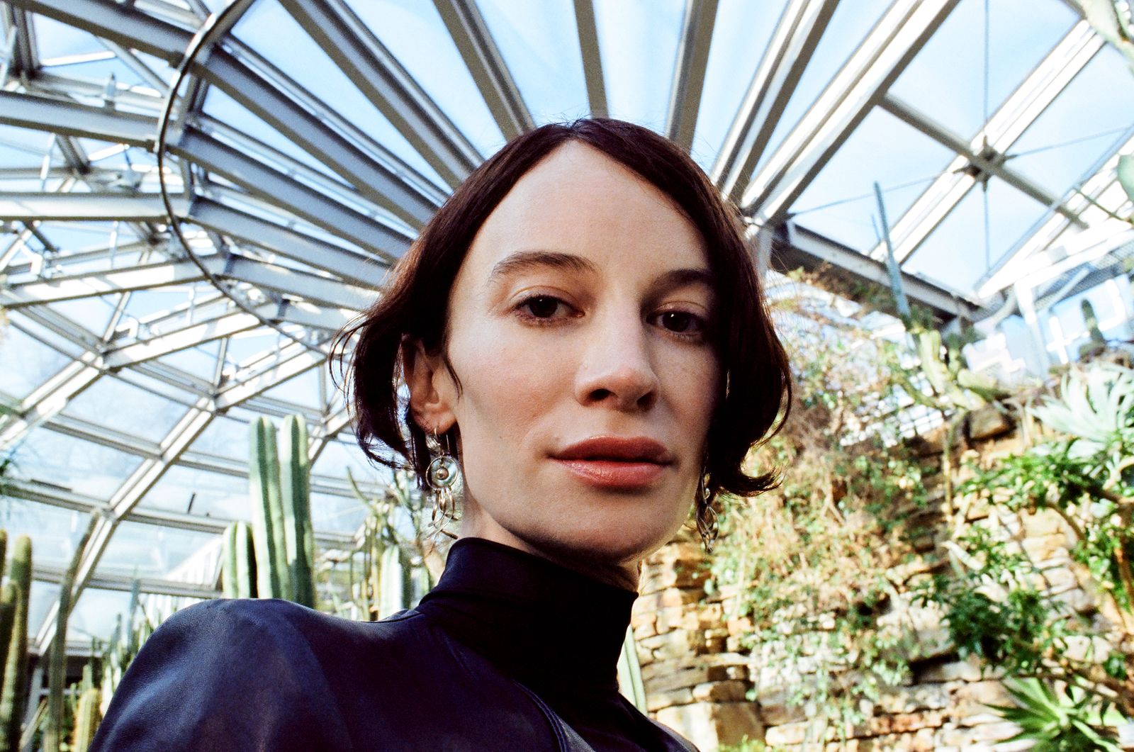 A closeup headshot portrait photograph of Alif Hilalm, who appears to be standing in an arboretum, with plants including cacti and a glass roof behind her
