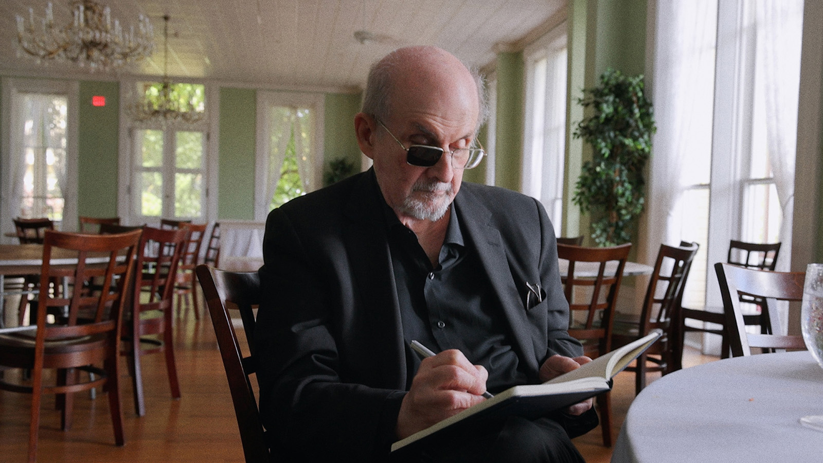A still image from Knife: The Attempted Murder of Salman Rushdie, showing the author writing in a notebook while sitting in what looks like a dining room