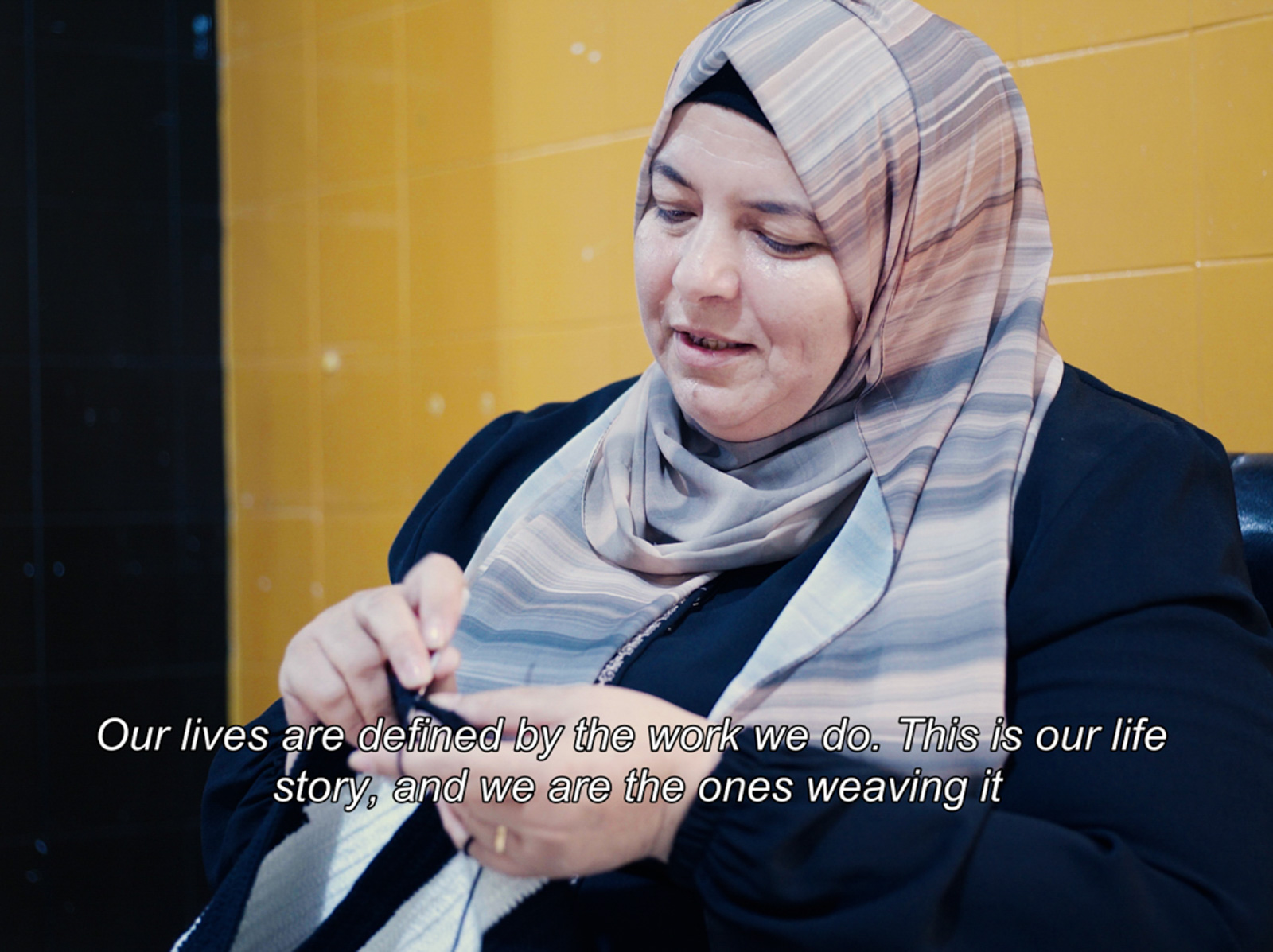 A still from a documentary project between Lamsa and David Gösta Dawson interviewing Syrian crochet artisans, showing a woman wearing a headscarf while hand weaving. The still has the quote caption: 'Our lives are defined by the work we do. This is our life story, and we are the ones weaving it.'