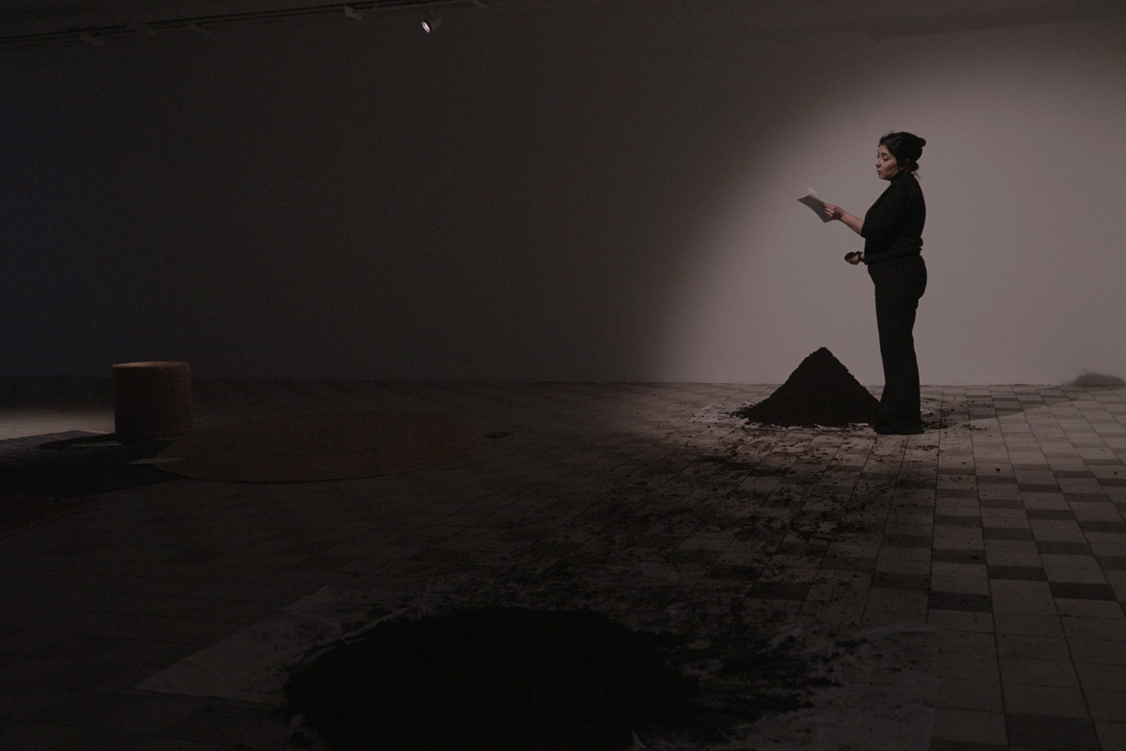 A photograph of Bahar Borna Faraz delivering a one-off performance about grief as part of Sacred Spaces. Faraz appears on the right of the image, the rest of which is shrouded in darkness, dressed all in black and standing beside what looks like a pile of ashes, reading from notes held in her left hand
