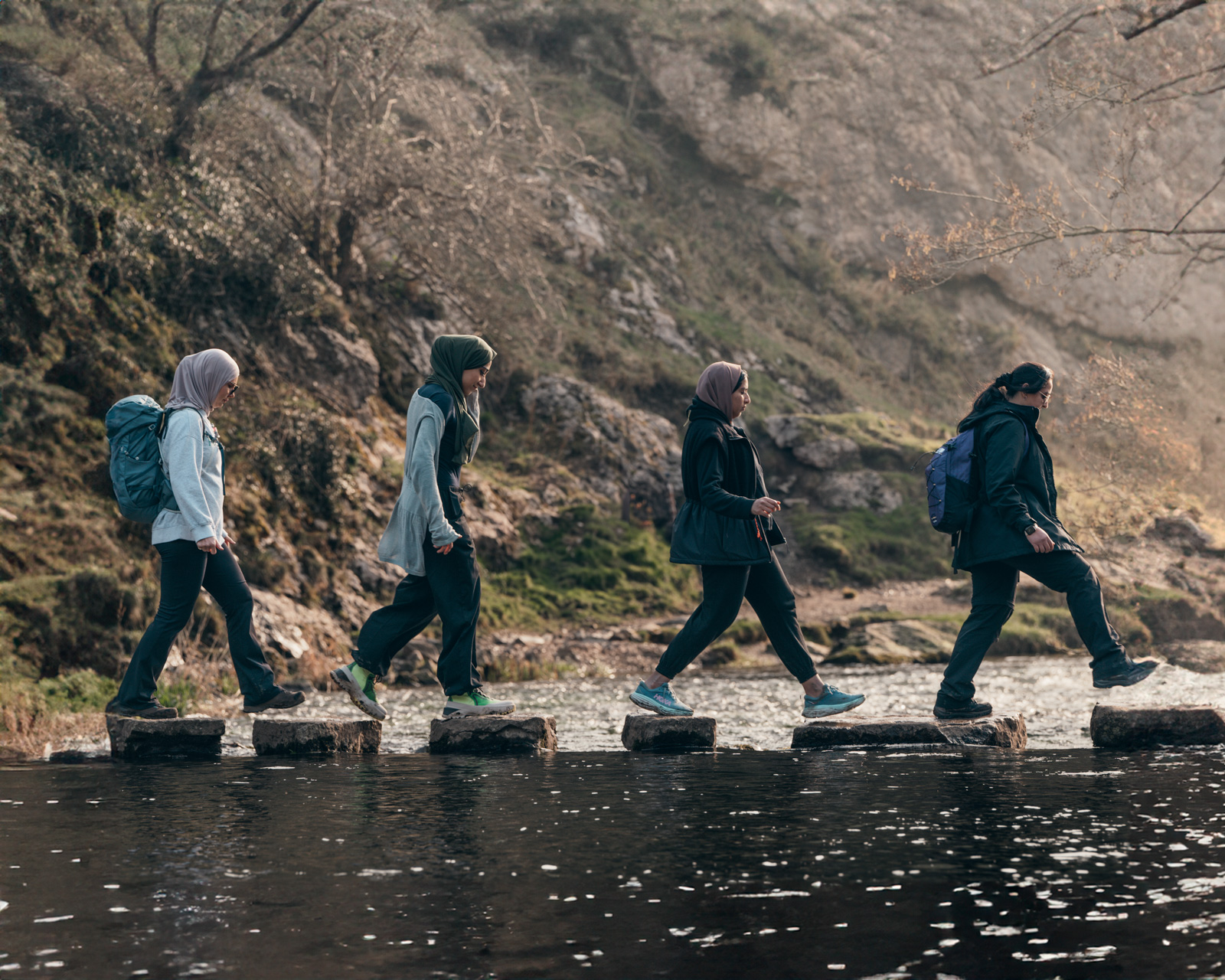 A photograph of four women crossing stepping stones in Dovedale in the Peak District during the Muslim Hikers Ramadan Retreat, March 2025