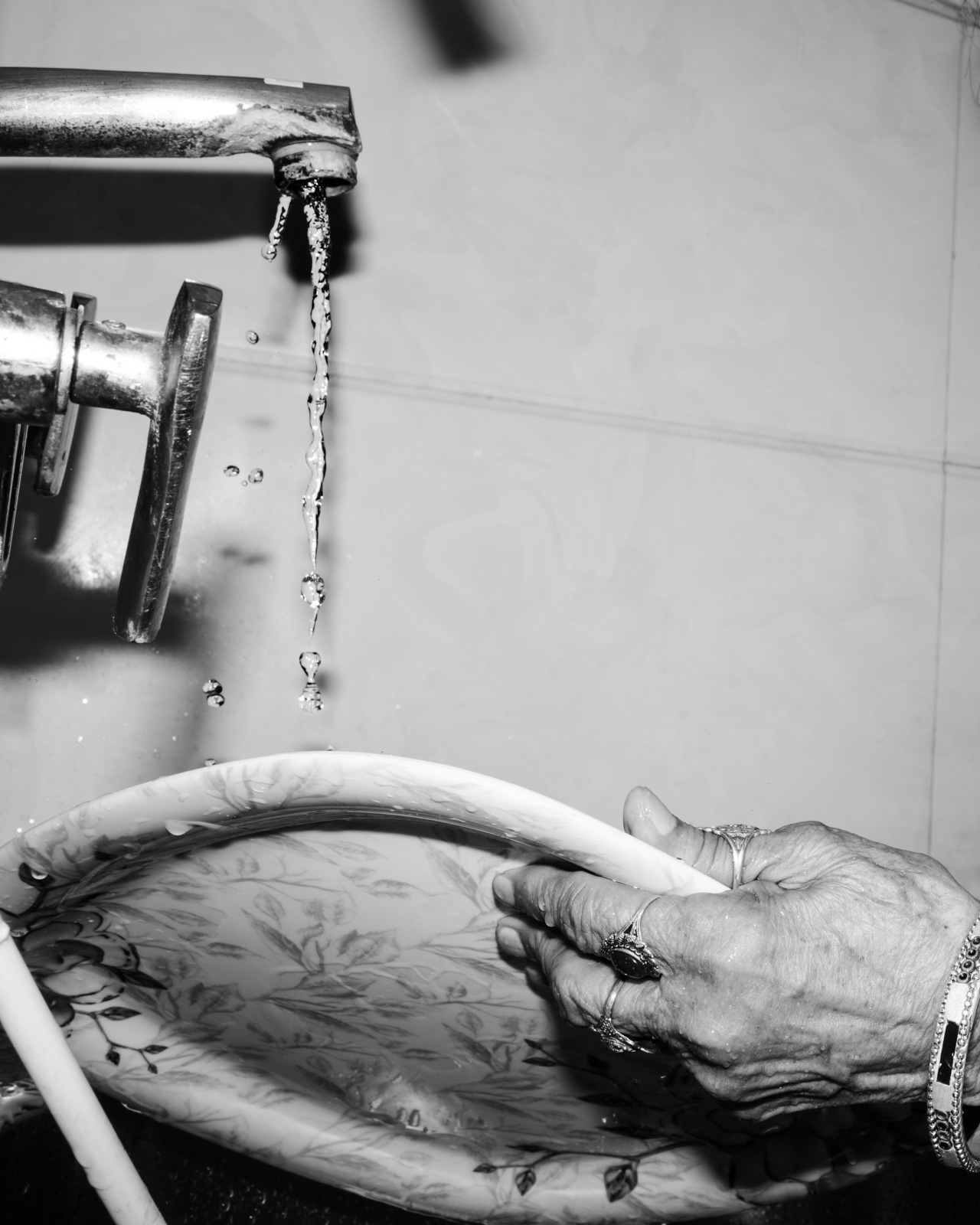 A black and white photograph from Aman Alam's series Ozymandias, documenting his grandmother's life with Alzheimer's, titled Cleaning the Dishes, which became an obsessive compulsion for her. The close-up image of tap water hitting a plate held by a hand adorned with rings and a bracelet shows Alam's mother cleaning dishes at 3am