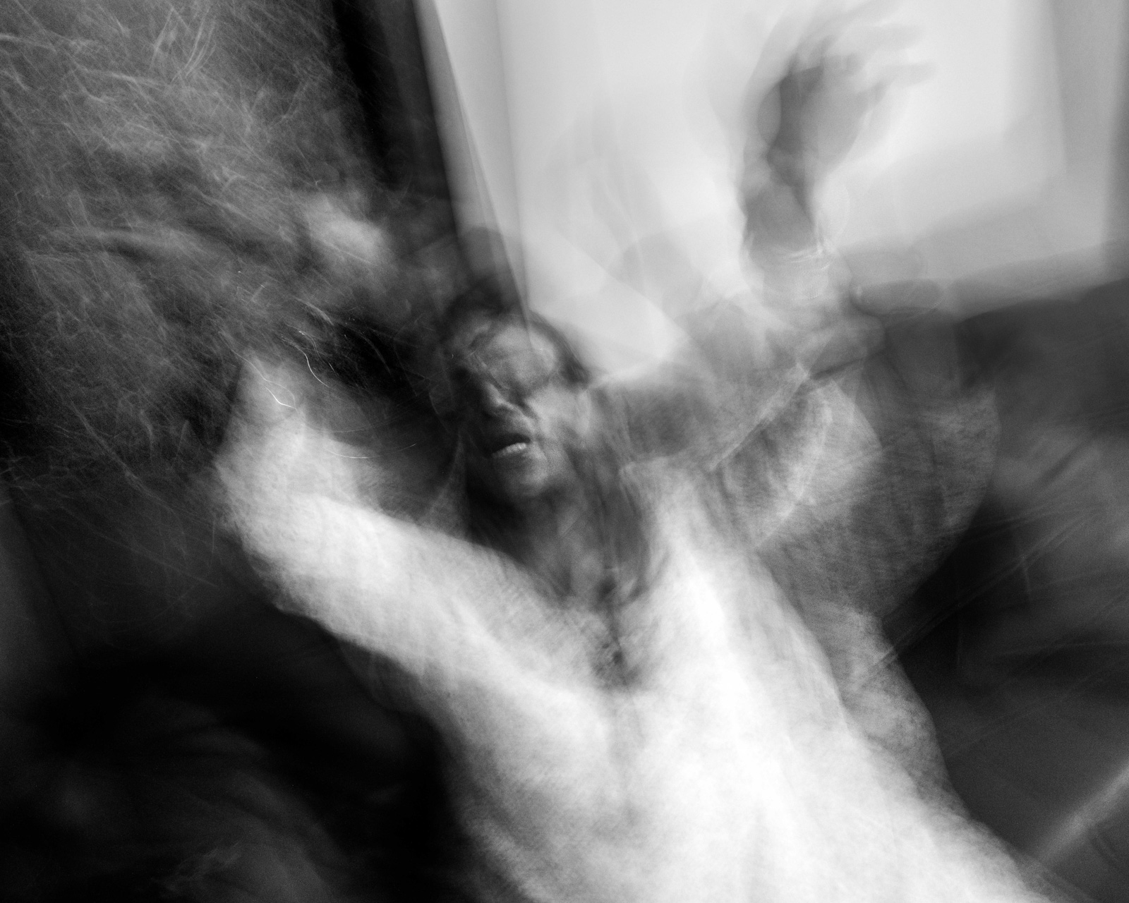 An out of focus black and white photograph from Aman Alam's series Ozymandias, documenting his grandmother's life with Alzheimer's, showing her seated, waving her arms in the air