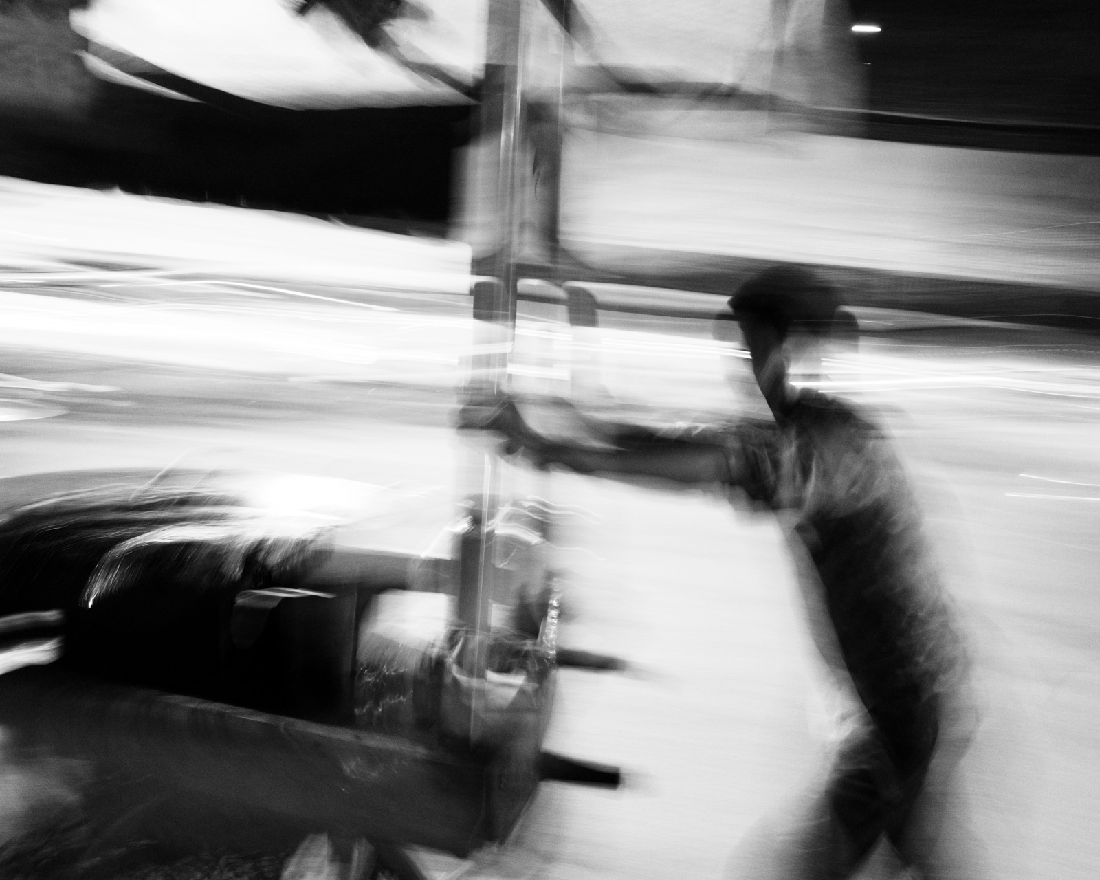 An out of focus black and white photograph from Aman Alam's series Ozymandias, documenting his grandmother's life with Alzheimer's, titled Agra Lunatic Asylum. The image shows a man pushing what appears to be a hospital gurney with a patient in it