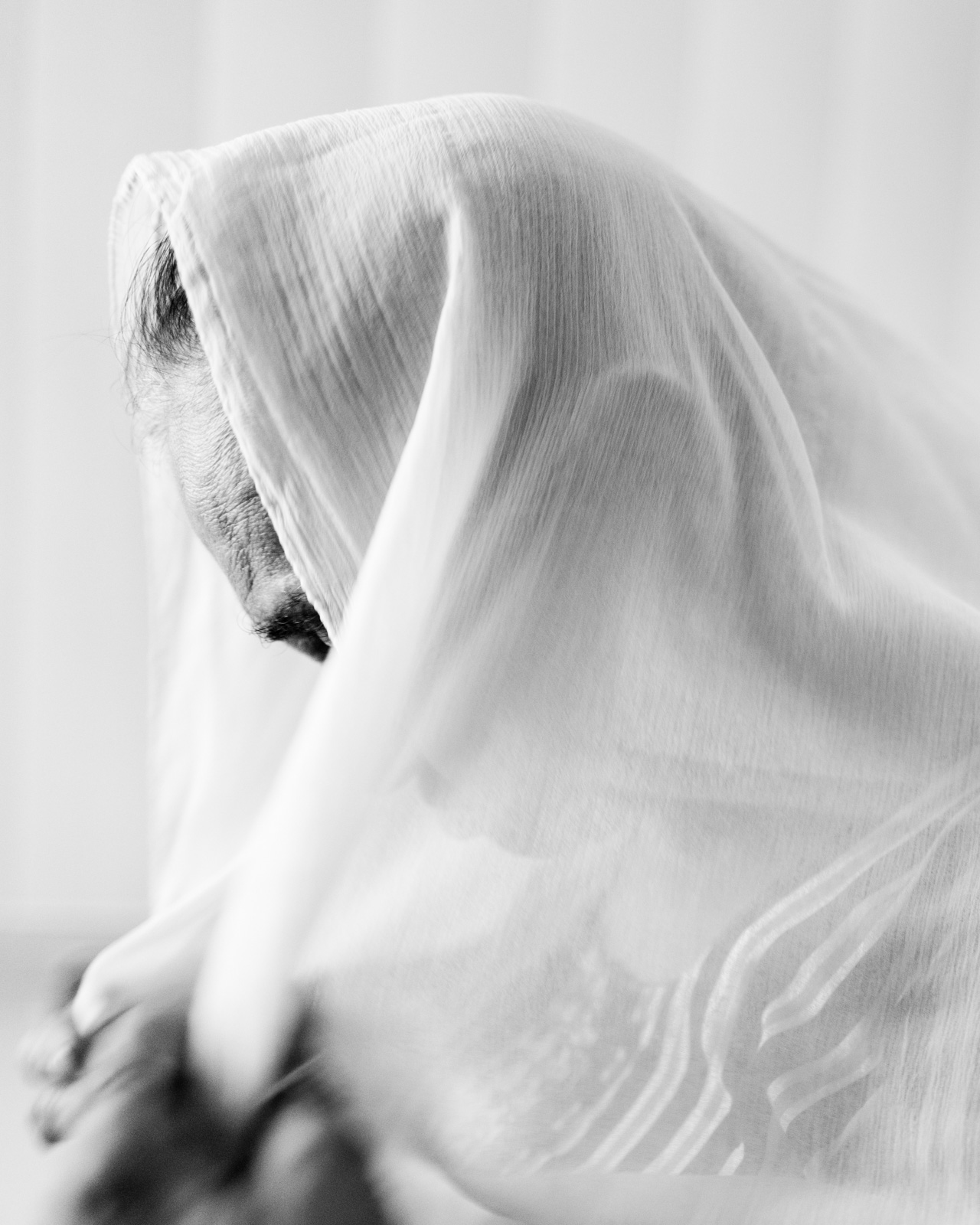 A black and white image of a woman with a white shawl over her head, from Aman Alam's series Ozymandias, documenting his grandmother's life with Alzheimer's, titled Naseem at prayer. Alam says that after forgetting almost everyone and everything around her, Naseem still remembered her prayers perfectly. 'She seems
confident and comfortable when she prays. In those moments I get glimpses of the woman who raised me and a reminder that she is still with me.'