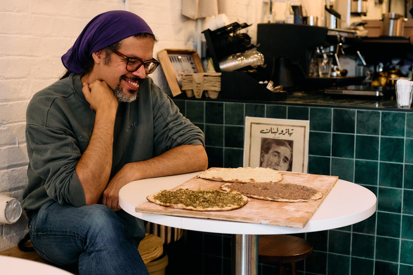 A photograph of Hyphen journalist Samir Jeraj smiling as he sits at a table in Edami, the east London Lebanese bakery, with labneh, minced beef and za'atar manakish on a wooden board in front of him