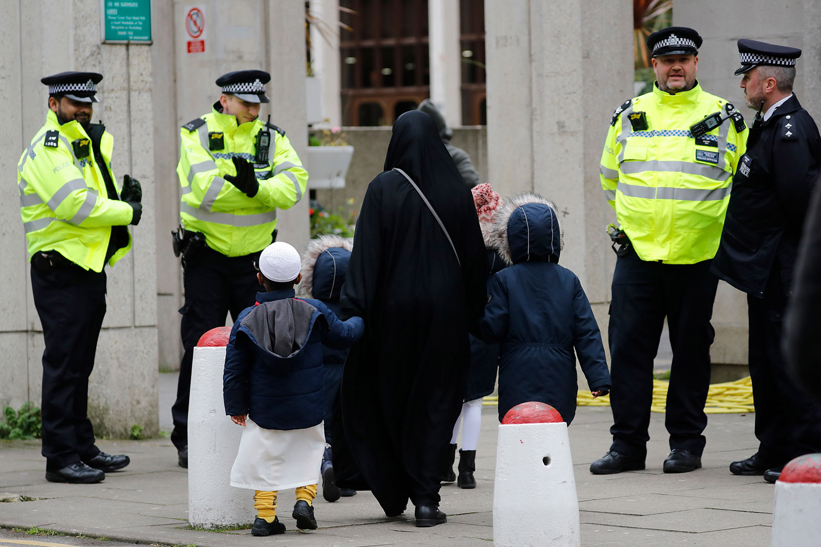 Three police in bright yellow jackets face the camera and flank a young family entering a building, who face away from us.