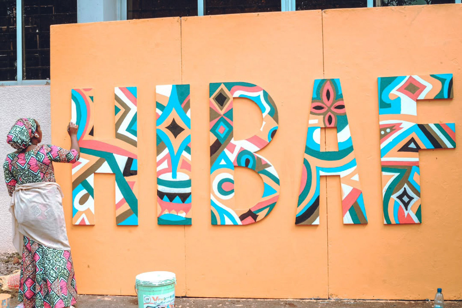 A photograph of a woman painting a large, brightly coloured 'HIBAF' (Hausa International Books and Arts Festival) sign