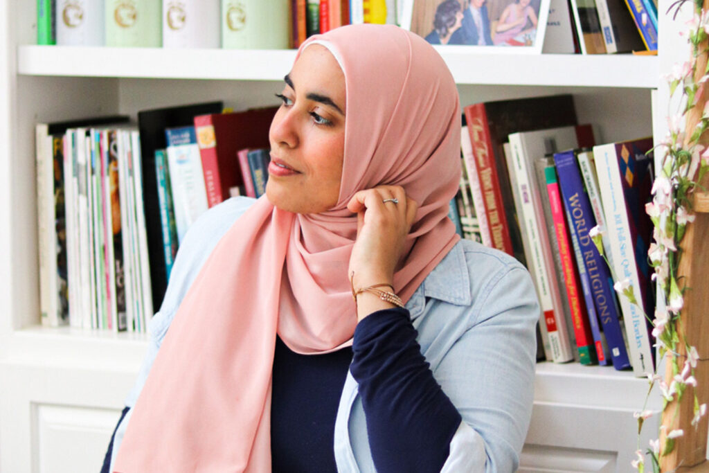 A photograph of author and cartoonist Huda Fahmy, wearing a salmon pink headscarf and sitting down in front of shelves of books