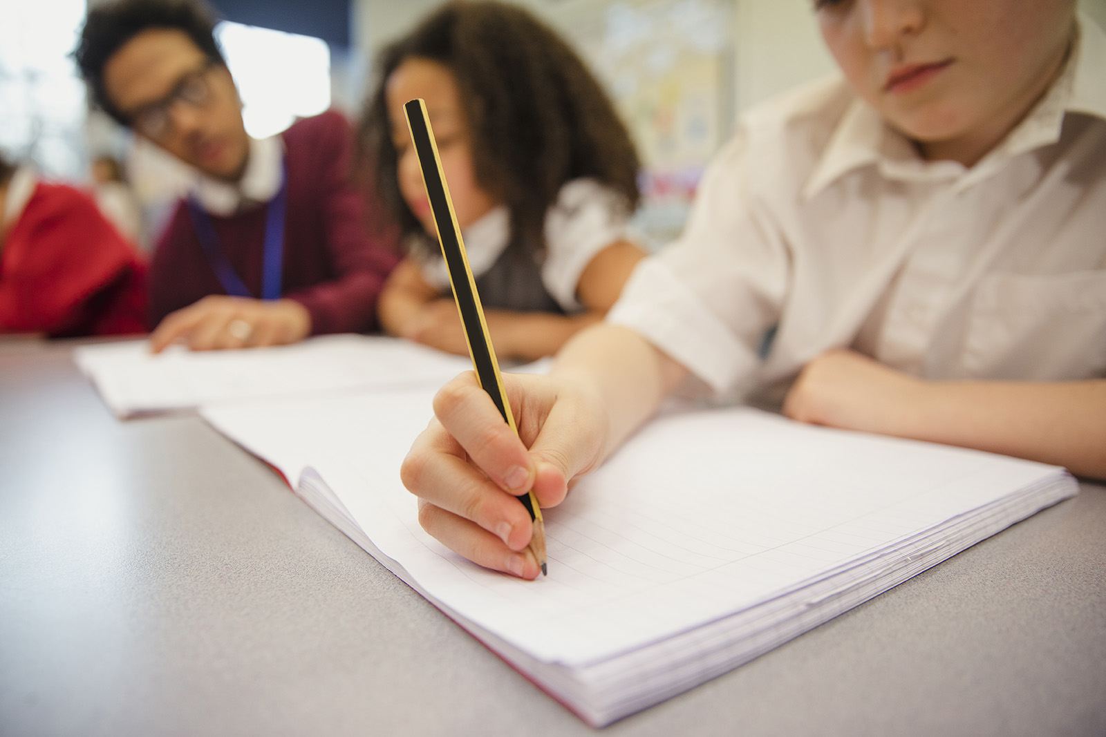A close-up of a young boy's hand writing in a notebook at a desk with other children in the background