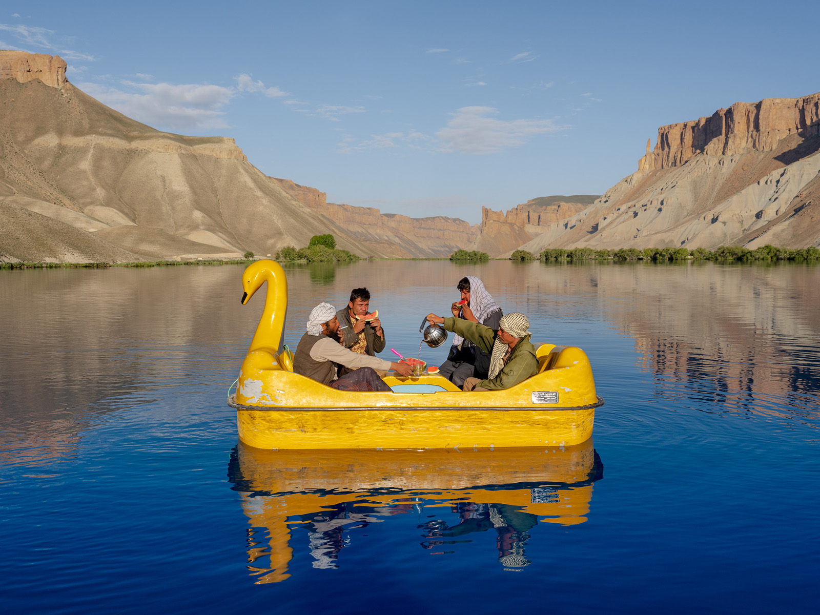 A photograph of four men sitting in a yellow, swan-shaped boat in a lake surrounded by arid cliffs and screes, drinking tea and eating watermelon, from Daniel Malikyar's new photobook Afghanistan