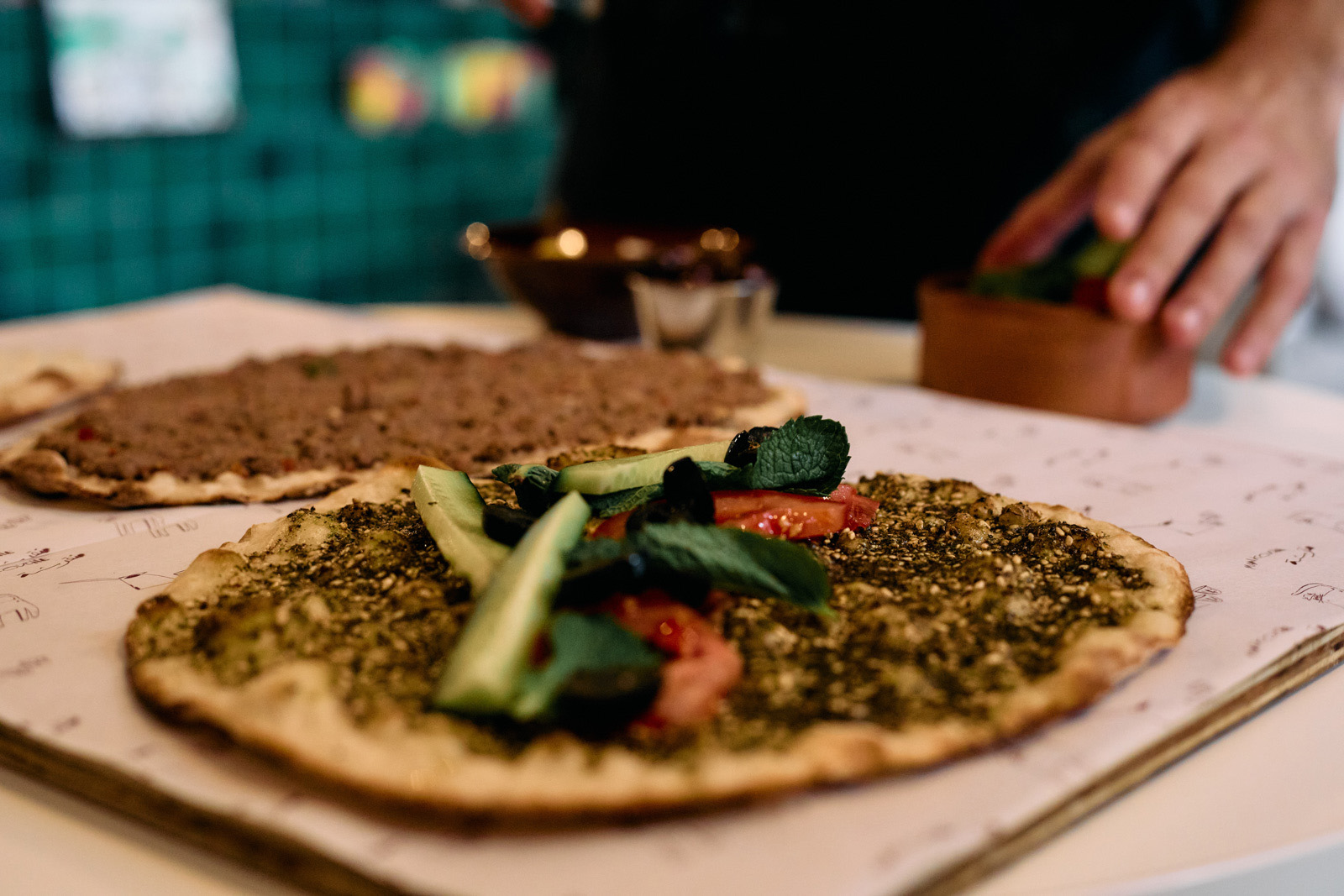 A close-up photograph of Edami's za'atar (on the right), garnished with cucumber, tomota and mint, and minced lamb manakish