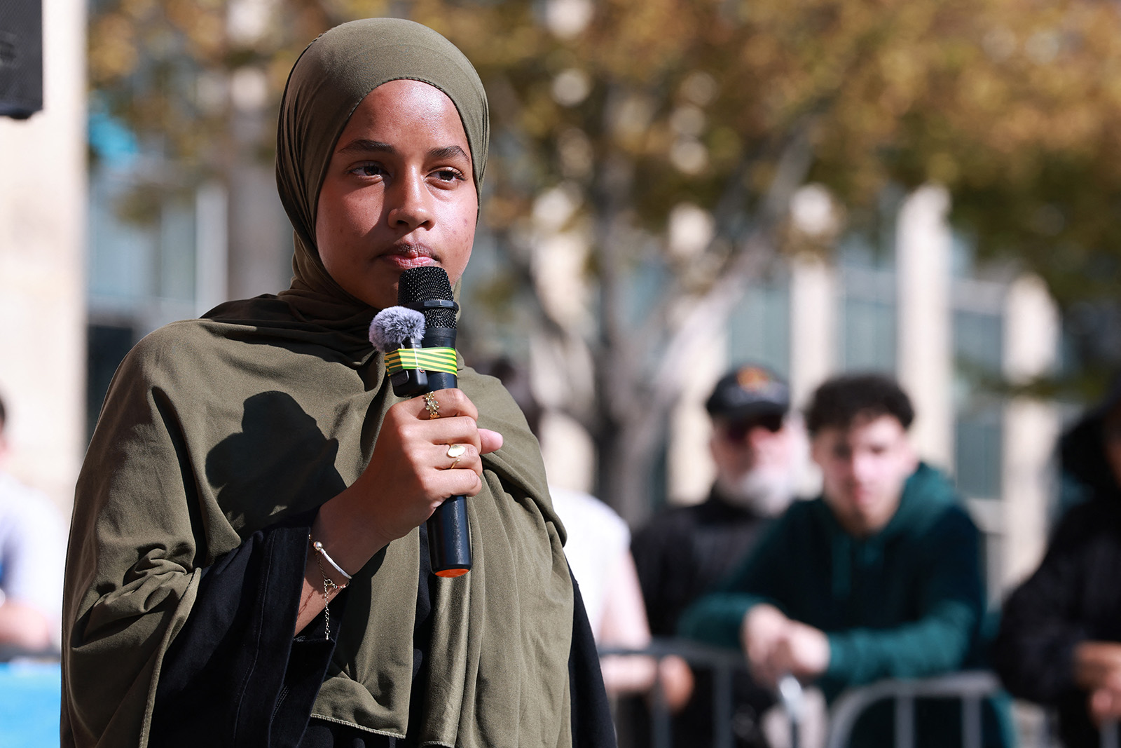 A young woman in a dark green abaya and hijab speaks into a microphone