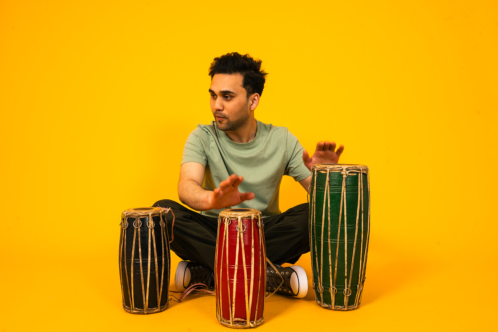 A photograph of Nepali percussionist Anmol Mohara, seated against a yellow background playing three hand drums