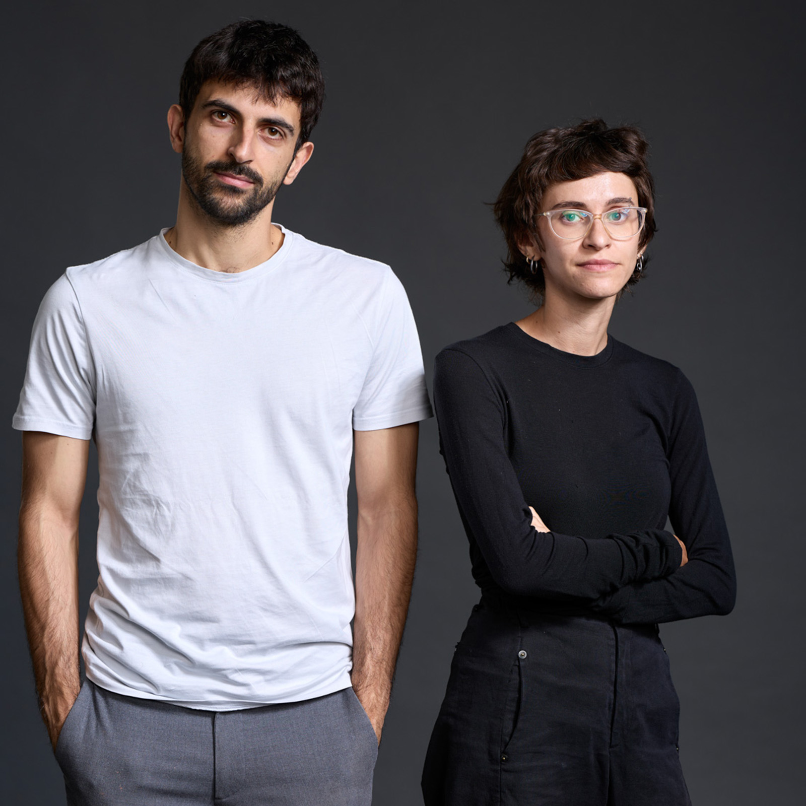 A portrait photograph of NAZA documentary directors Yuval Abraham and Rachel Szor, standing side by side with Abraham, on the left, wearing a white T-shirt and grey trousers; and Szor wearing a black long-sleeved top and trousers