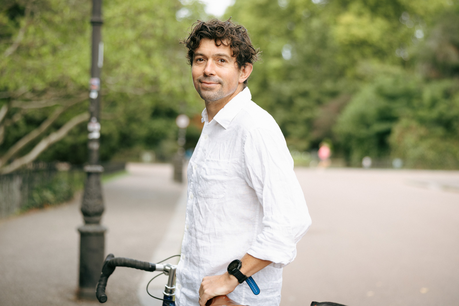 A posed side-on photograph of Dermot Bryers in Battersea Park, smiling and wearing a white shirt as he leans against his bike