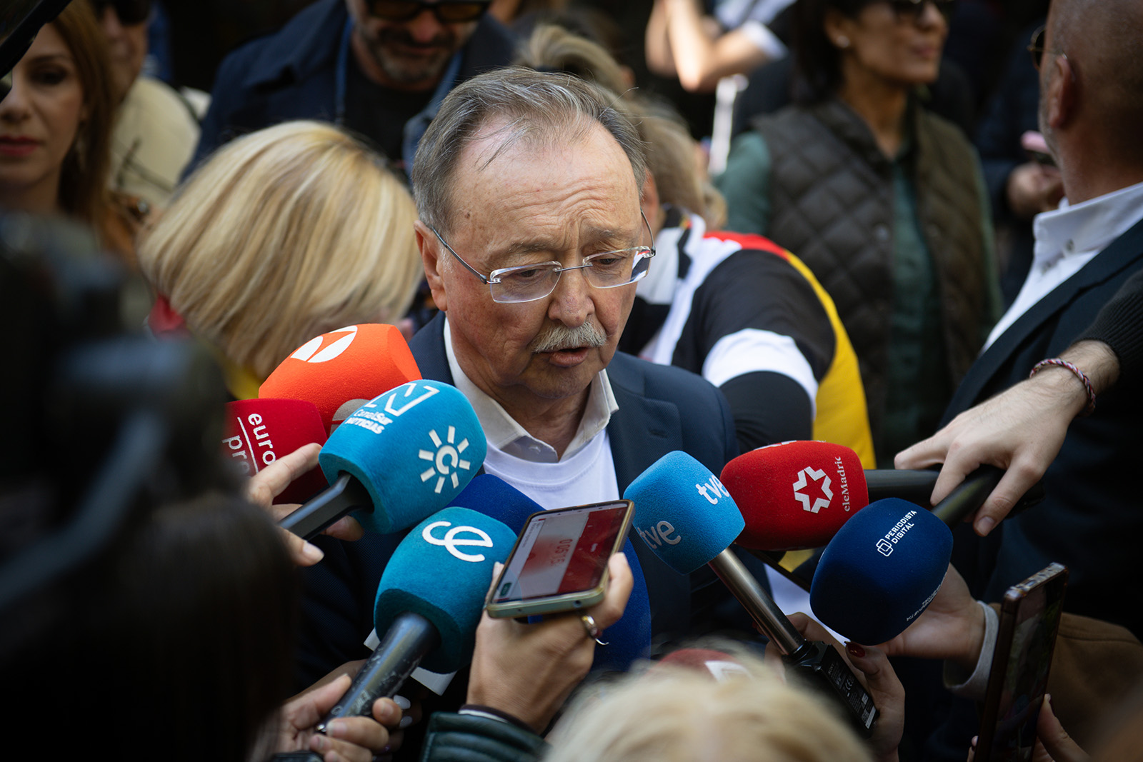 A man in glasses with a moustache speaks as microphones cluster towards him