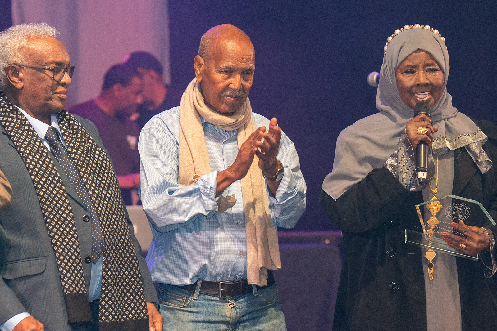 A photograph featuring novelist Nuruddin Farah, on stage flanked by a man and a woman, during the Somali Week Festival 2025. This year's event will celebrate his 80th birthday