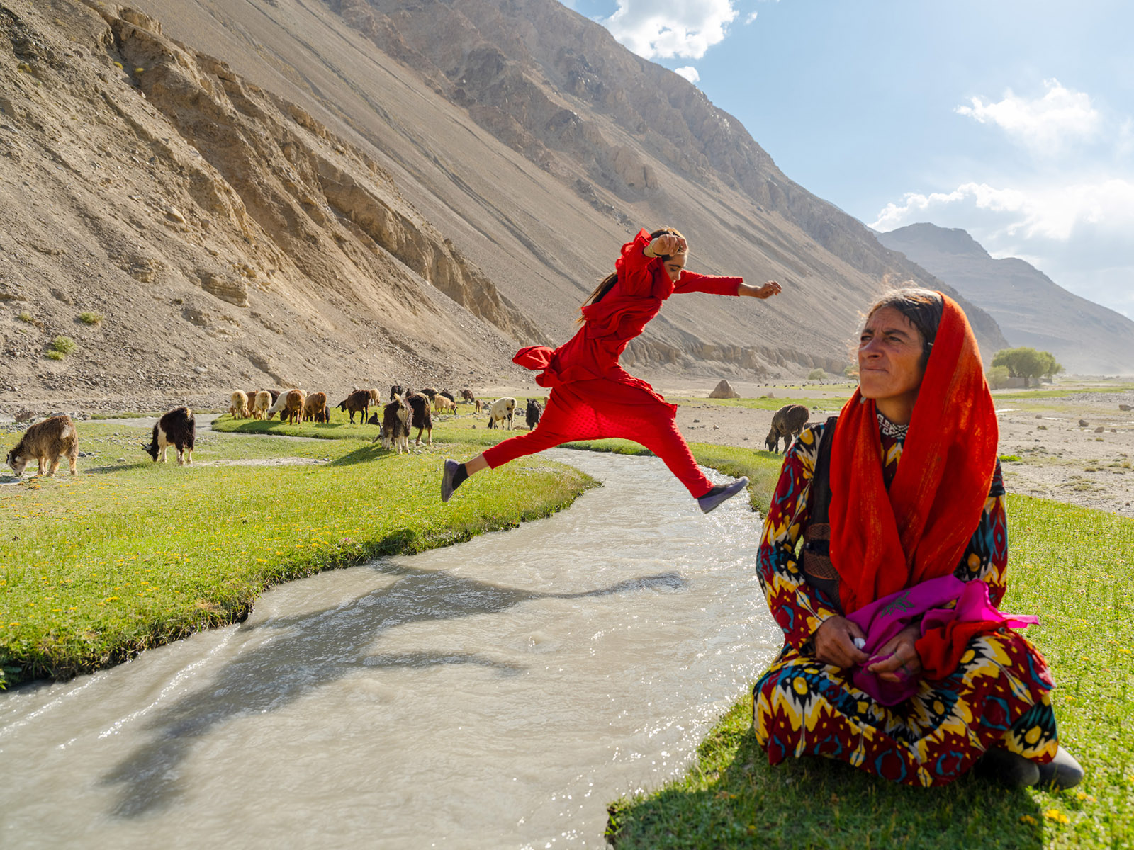 A photograph of a girl leaping over a stream, with cattle grazing in the background a woman seated in the foreground, from Daniel Malikyar's new photobook Afghanistan
