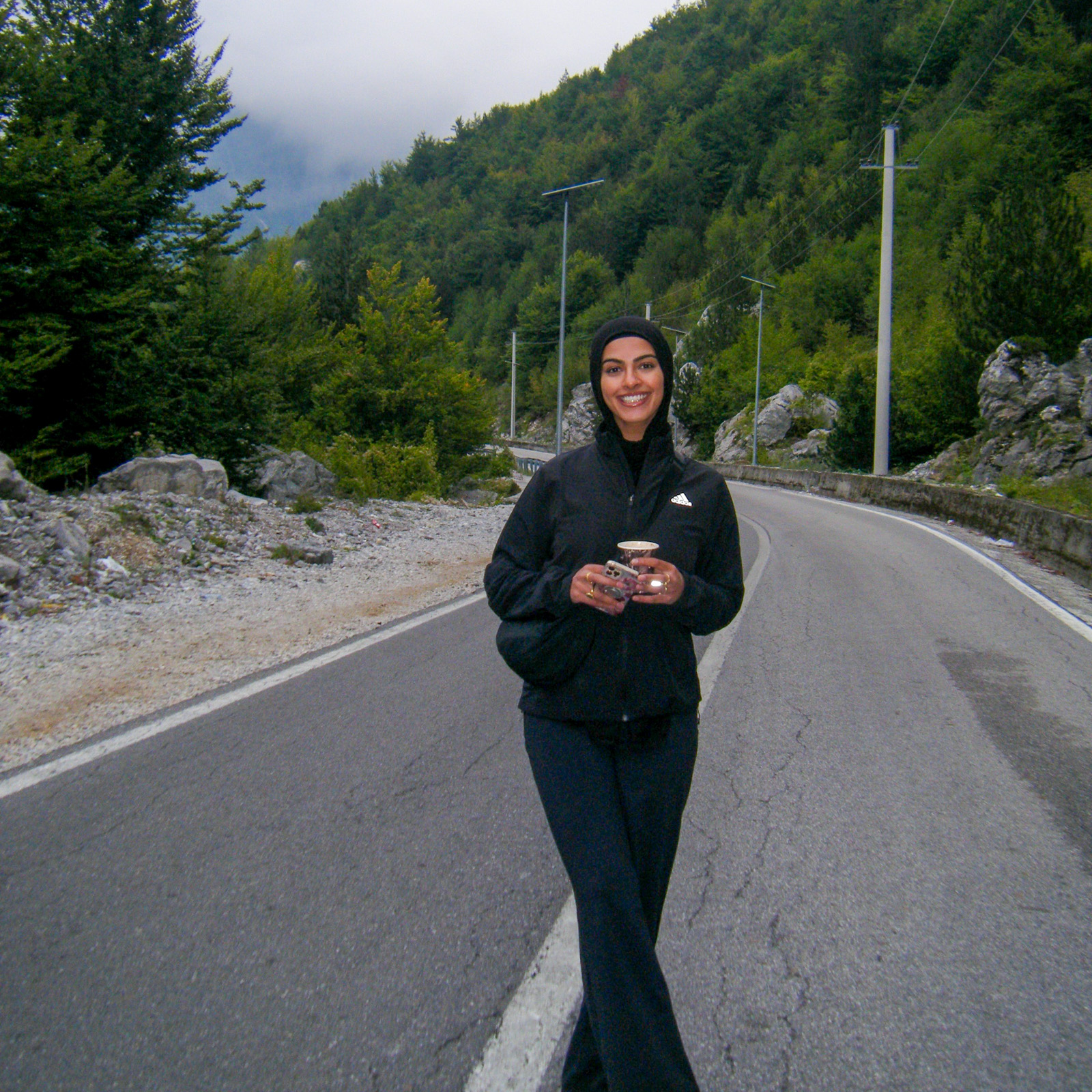 A photograph of Aisha Hussain, smiling for the camera while standing in the middle of a country road holding her phone and a drink in her hands