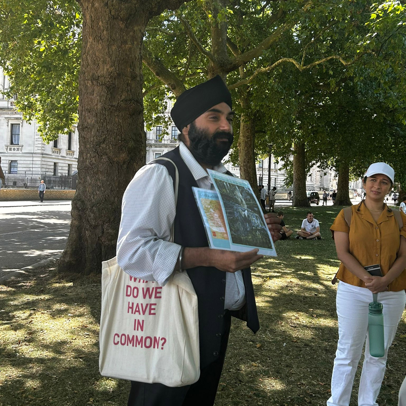 A man in a turban and a shirt holds up a black and white poster while a tourist looks on. His audience is cropped out of the image. He has a bag saying 'what do we have in common?' on it