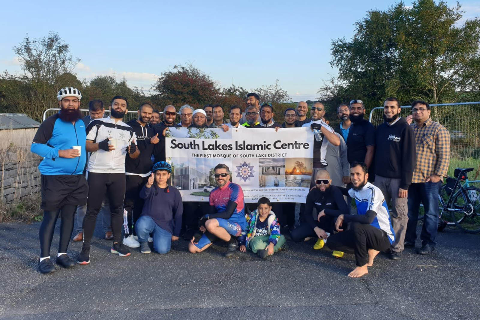 A photograph showing Slow Spokes cycle club members with a South Lakes Islamic Centre banner, proclaiming it to be 'the first mosque of South Lake District'