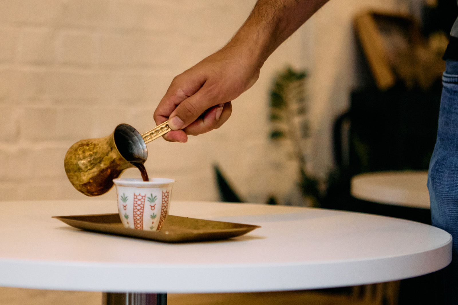A close-up photograph of someone pouring Levantine-style coffee from a long-handled rakweh pot into a small cup at Edami